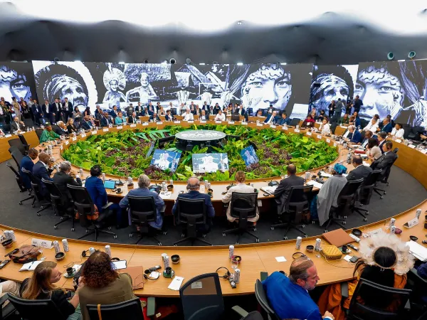 Blick in den Verhanldungsraum der COP30. Zwei Tischkreise, in der Mitte tropische Pflanzen. An den Wänden hängen schwarz-weiß-Fotos von Indigenen.